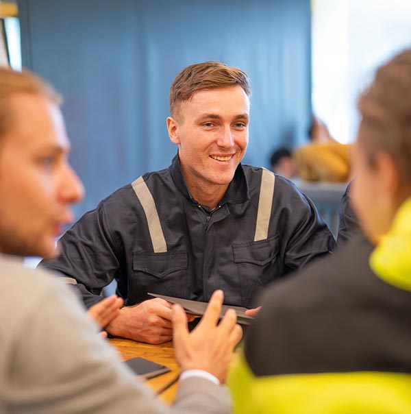 team of oil refinery engineers. wearing in safety wear, one man in casual wear, and another man in workwear discuss something at a table with a laptop. Business meeting, teamwork, and communication concept.